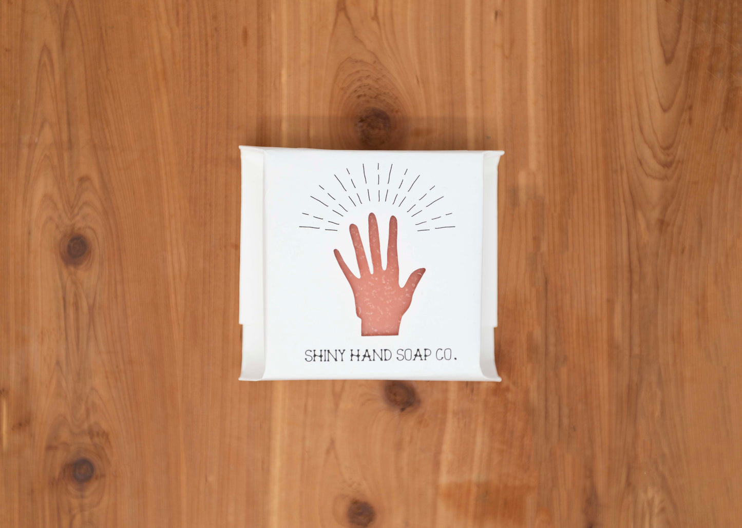 One vibrant coral peach colored geranium citrus soap bar wrapped in a white recycled paper with a hand cutout shape against a wooden cedar background.
