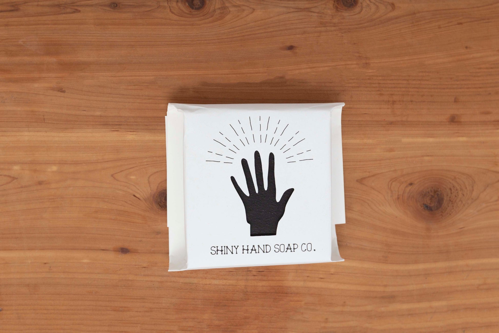 A charcoal black midnight anise soap bar wrapped in a crisp white recycled paper wrapper with a hand cutout rests on a warm brown cedar background