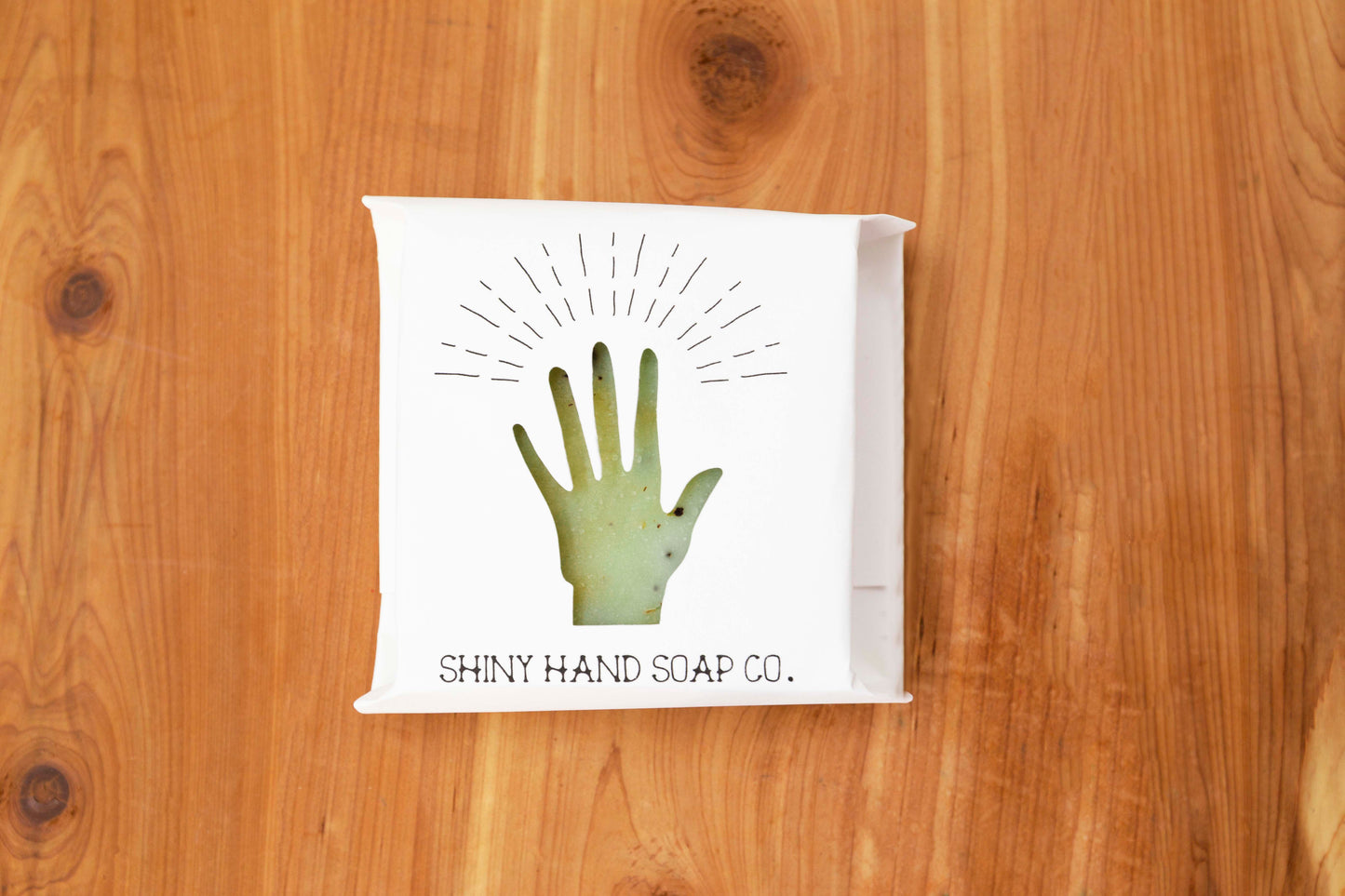 Pale Green Basil Mint Soap Bar flecked with bits of crushed coffee beans wrapped in a white recycled paper with a hand cutout shape against a wooden cedar background.