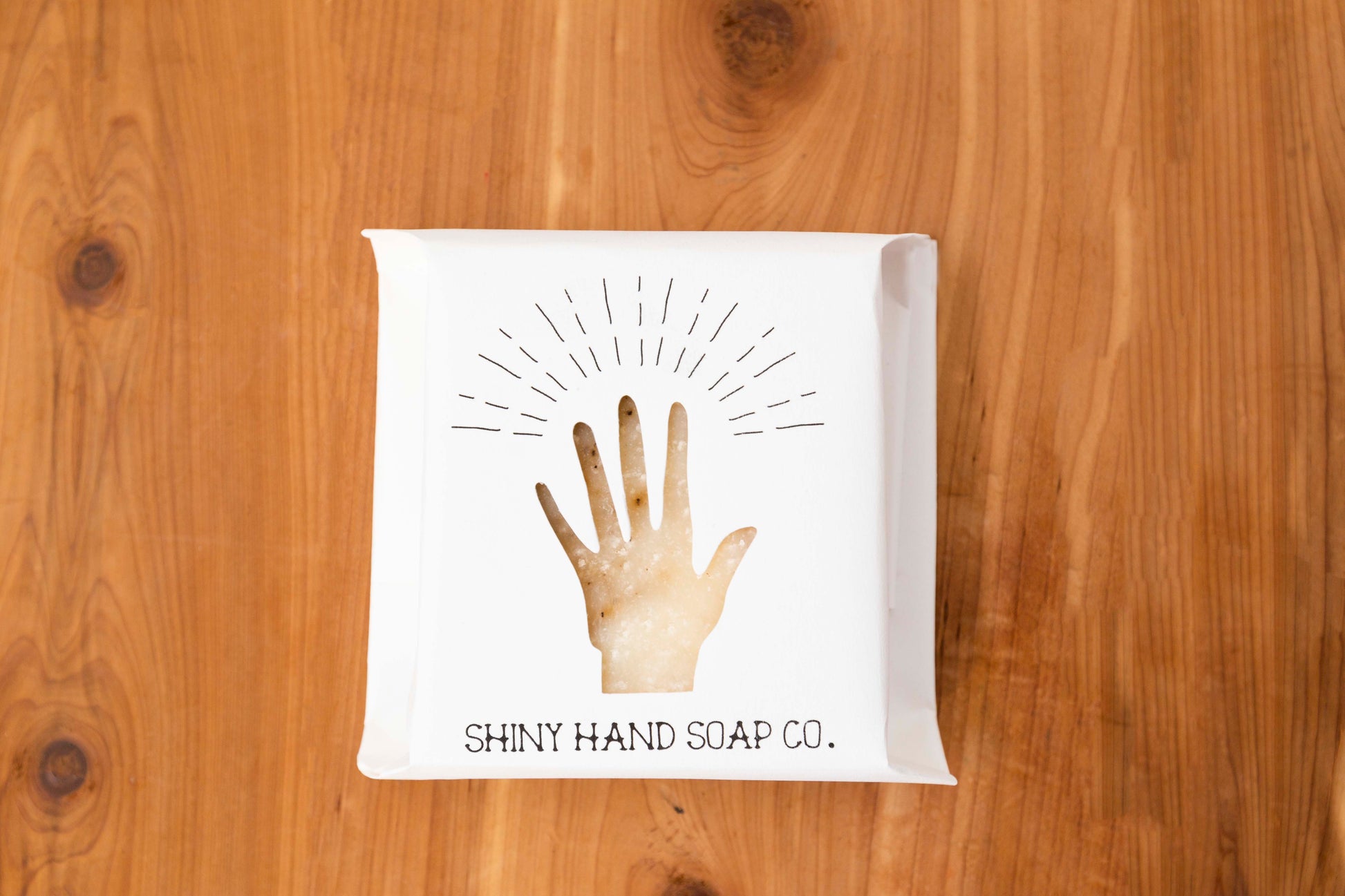 Ivory colored grapefruit black pepper soap bar with tiny specks of a black pepper swirl wrapped in a white recycled paper with a hand shaped cutout against a wooden cedar background.