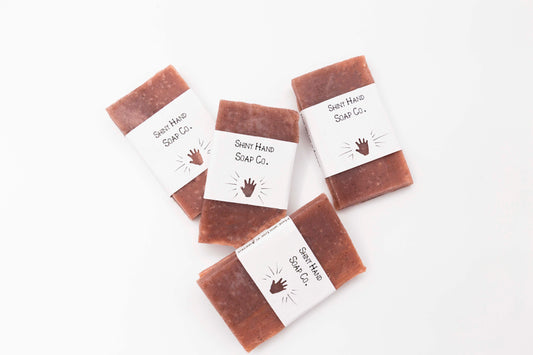 A miniature pale red Cinnamon rosemary soap bar speckled with dark brown cinnamon sits on a clean white background wrapped in a small paper wrapper with a hand cut out of it.