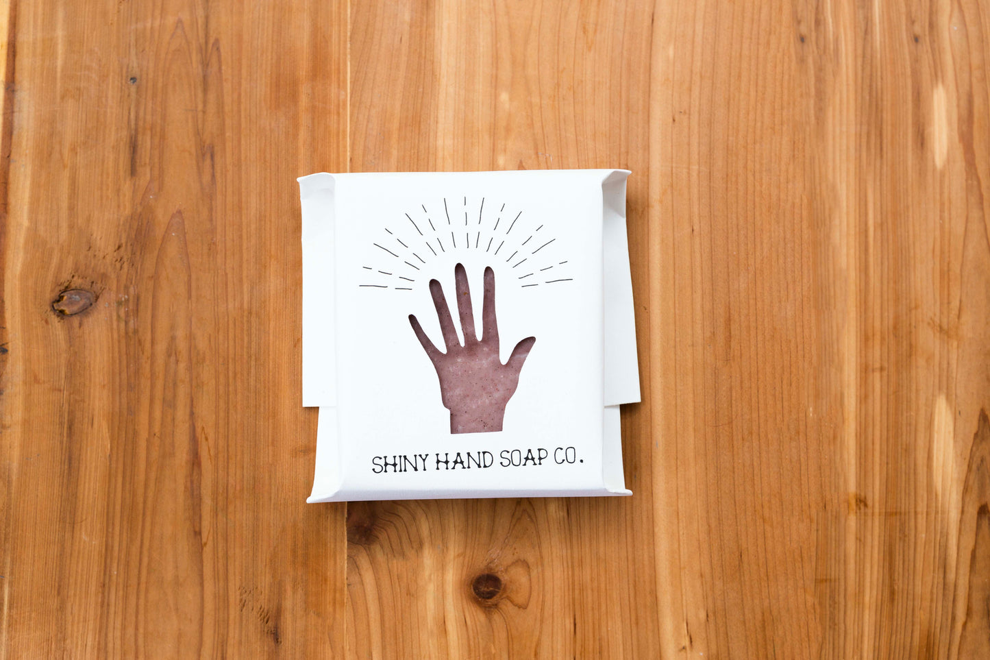 Pale red Cinnamon rosemary soap bar speckled with dark brown cinnamon wrapped in a white recycled paper with a hand shaped cutout against a wooden cedar background.