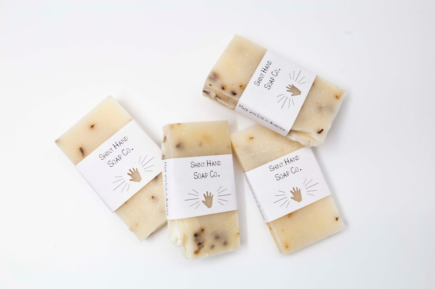 A miniature ivory colored lavender blossom soap bar speckled with pale purple lavender flowers sits on a clean white background wrapped in a small paper wrapper with a hand cut out of it.
