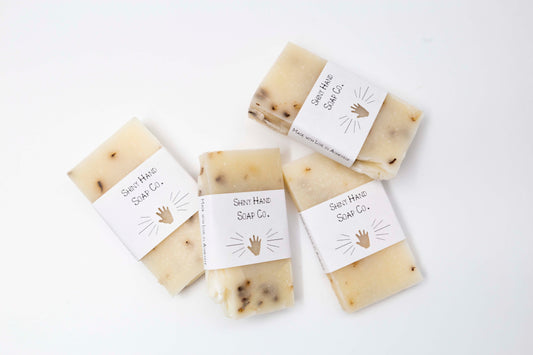 A miniature ivory colored lavender blossom soap bar speckled with pale purple lavender flowers sits on a clean white background wrapped in a small paper wrapper with a hand cut out of it.