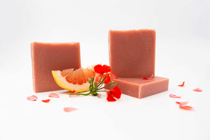 Three vibrant coral peach colored geranium citrus soap bars sit on a clean white background surrounded by bright red geranium flowers, fresh flower petals, and fresh pink and yellow grapefruit. 