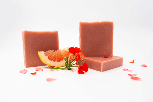 Three vibrant coral peach colored geranium citrus soap bars sit on a clean white background surrounded by bright red geranium flowers, fresh flower petals, and fresh pink and yellow grapefruit.