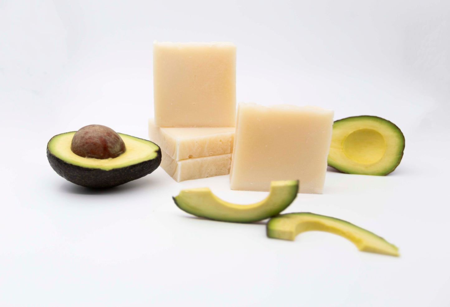 Four creamy ivory white bars of Avocado Butter bar soap are stacked on a clean white background with fresh bright green avocados surrounding them