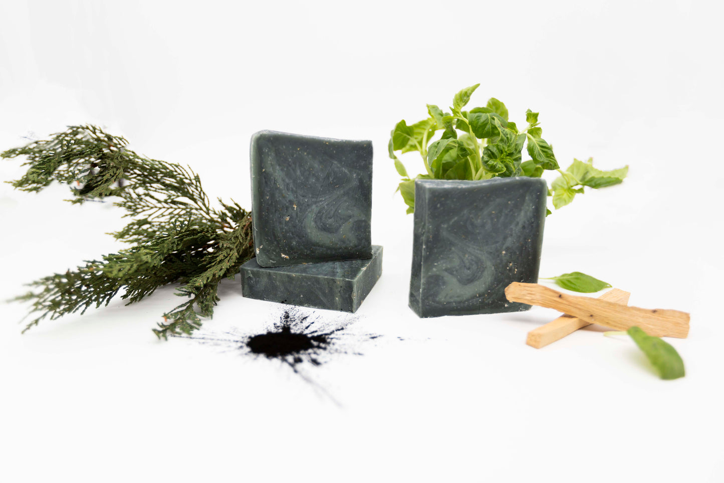 Hunter dark green ancient wood bar soaps with a dark grey marbled swirl pattern flecked with ivory colored organic oats sit on a clean white background with a splattered burst of coal-black activated charcoal powder, light tan pieces of Palo Santo holy wood, deep green fresh cedar branches and organic bright green fresh basil leaves.