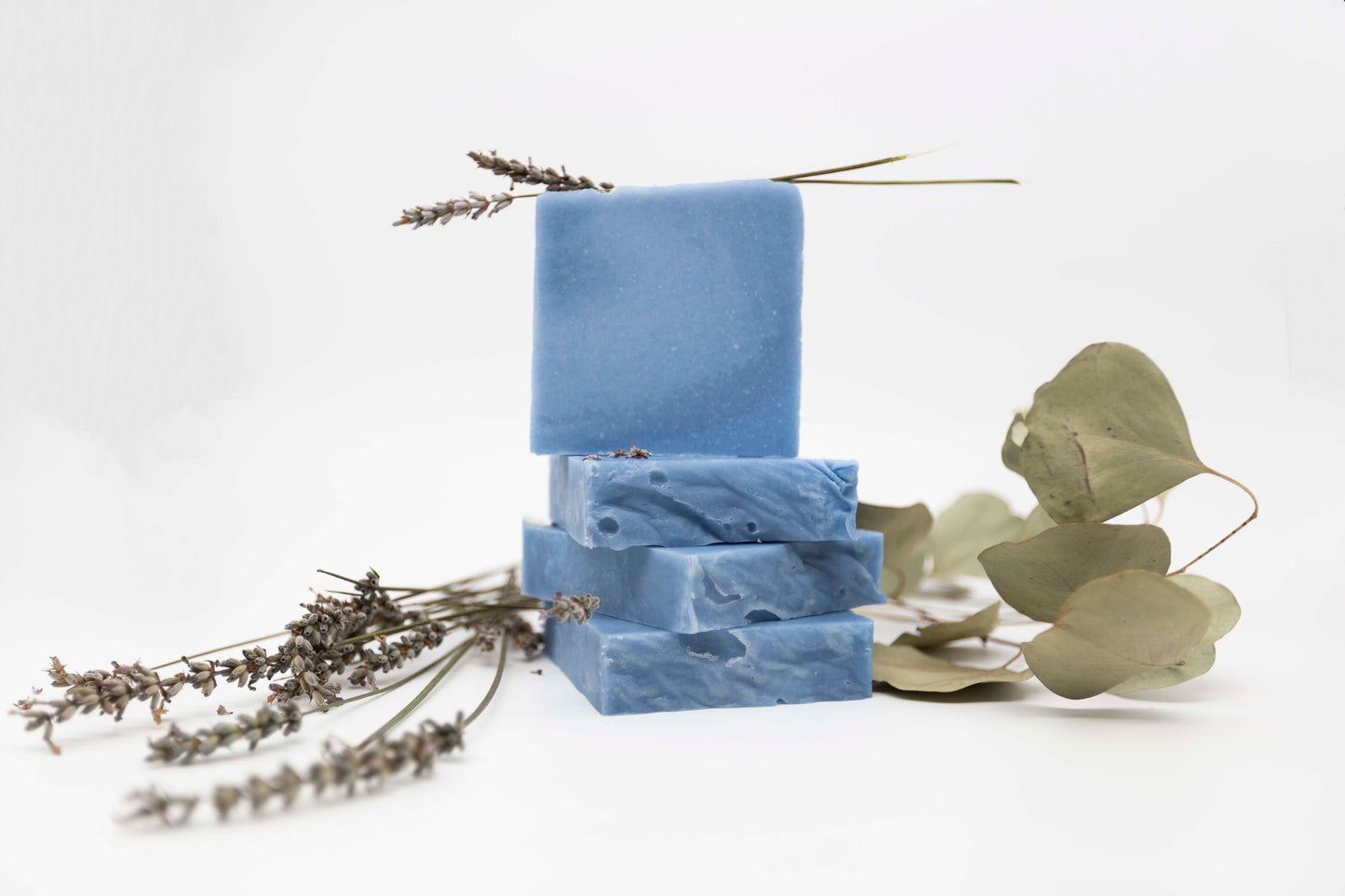 Four Cornflower blue bars of soap are stacked on a clean white background with real eucalyptus leaves and lavender flowers surrounding them