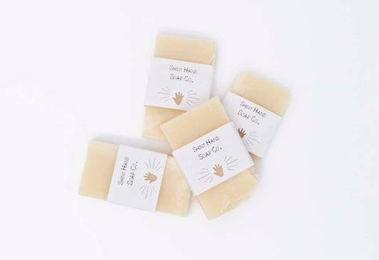 Four creamy ivory white miniature bars of Avocado Butter bar soap sit on a clean white background with white paper wrappers that have a hand shape cut out of them.