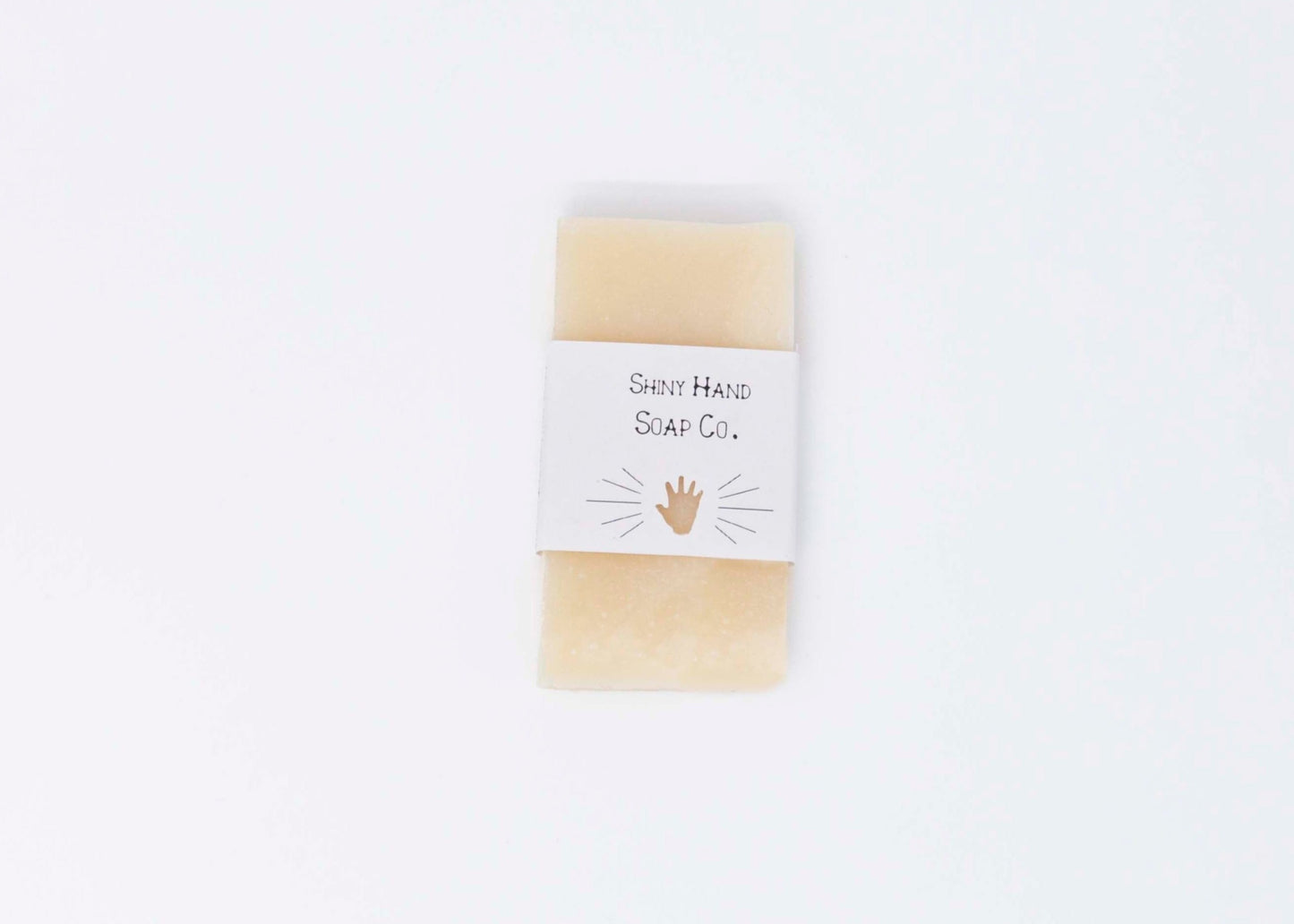 One creamy ivory white miniature bar of Avocado Butter bar soap sits on a clean white background with a white paper wrapper that has a hand shape cut out of it.