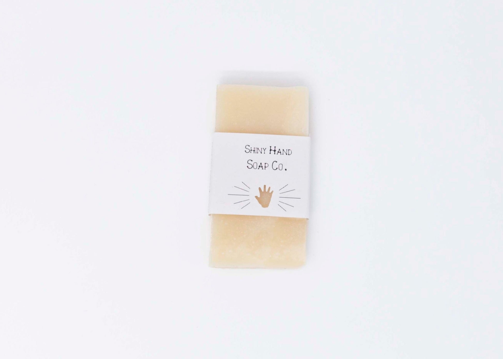 One creamy ivory white miniature bar of Avocado Butter bar soap sits on a clean white background with a white paper wrapper that has a hand shape cut out of it.