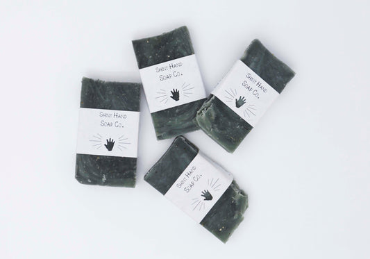 Four miniature Hunter dark green ancient wood bar soaps with a dark grey marbled swirl pattern flecked with ivory colored organic oats sit on a clean white background with white paper wrappers that have a hand shape cut out of them.