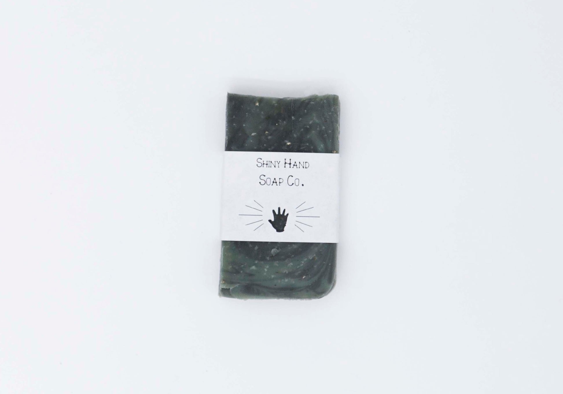 One miniature Hunter dark green ancient wood bar soap with a dark grey marbled swirl pattern flecked with ivory colored organic oats sits on a clean white background with white paper wrapper that has a hand shape cut out of it.