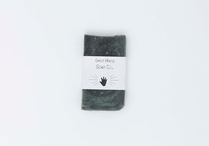 One miniature Hunter dark green ancient wood bar soap with a dark grey marbled swirl pattern flecked with ivory colored organic oats sits on a clean white background with white paper wrapper that has a hand shape cut out of it.