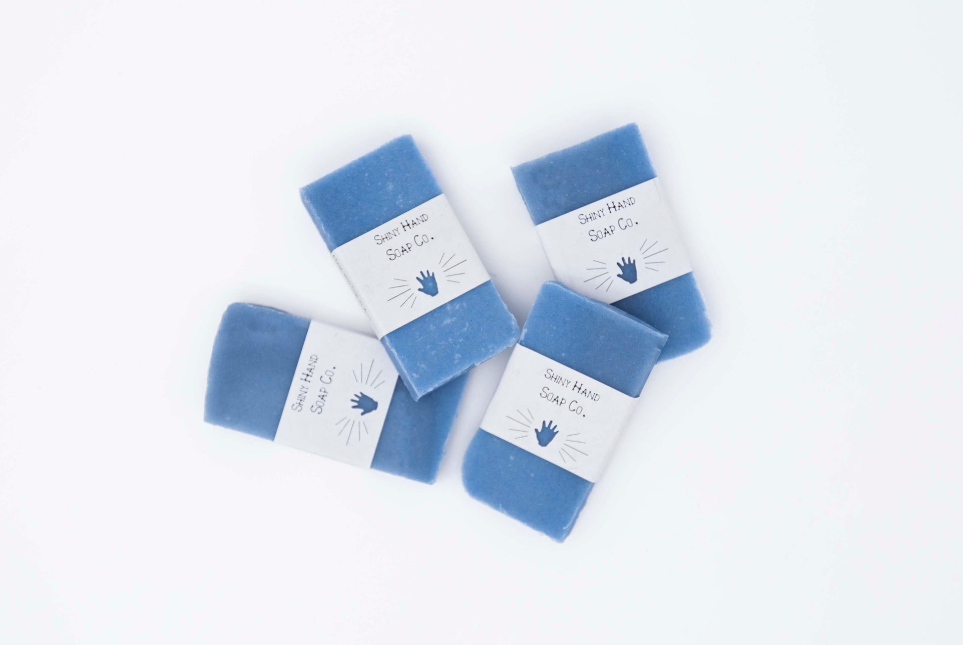 Four bright royal blue miniature bars of eucalyptus lavender bar soap sit on a clean white background with white paper wrappers that have a hand shape cut out of them.