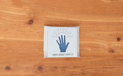 Royal cornflower blue eucalyptus soap bar  wrapped in a white recycled paper with a hand shaped cutout against a wooden cedar background.