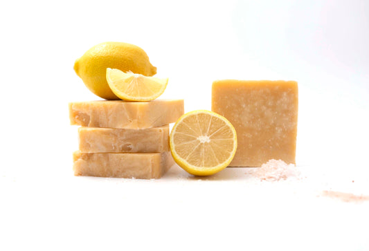 Rich yellow Lemon Sea Salt Soaps flecked with white salt crystals in front of a clean white backdrop with fresh juicy bright yellow lemons, white crystals of natural sea salt and pink flaky himalayan salt