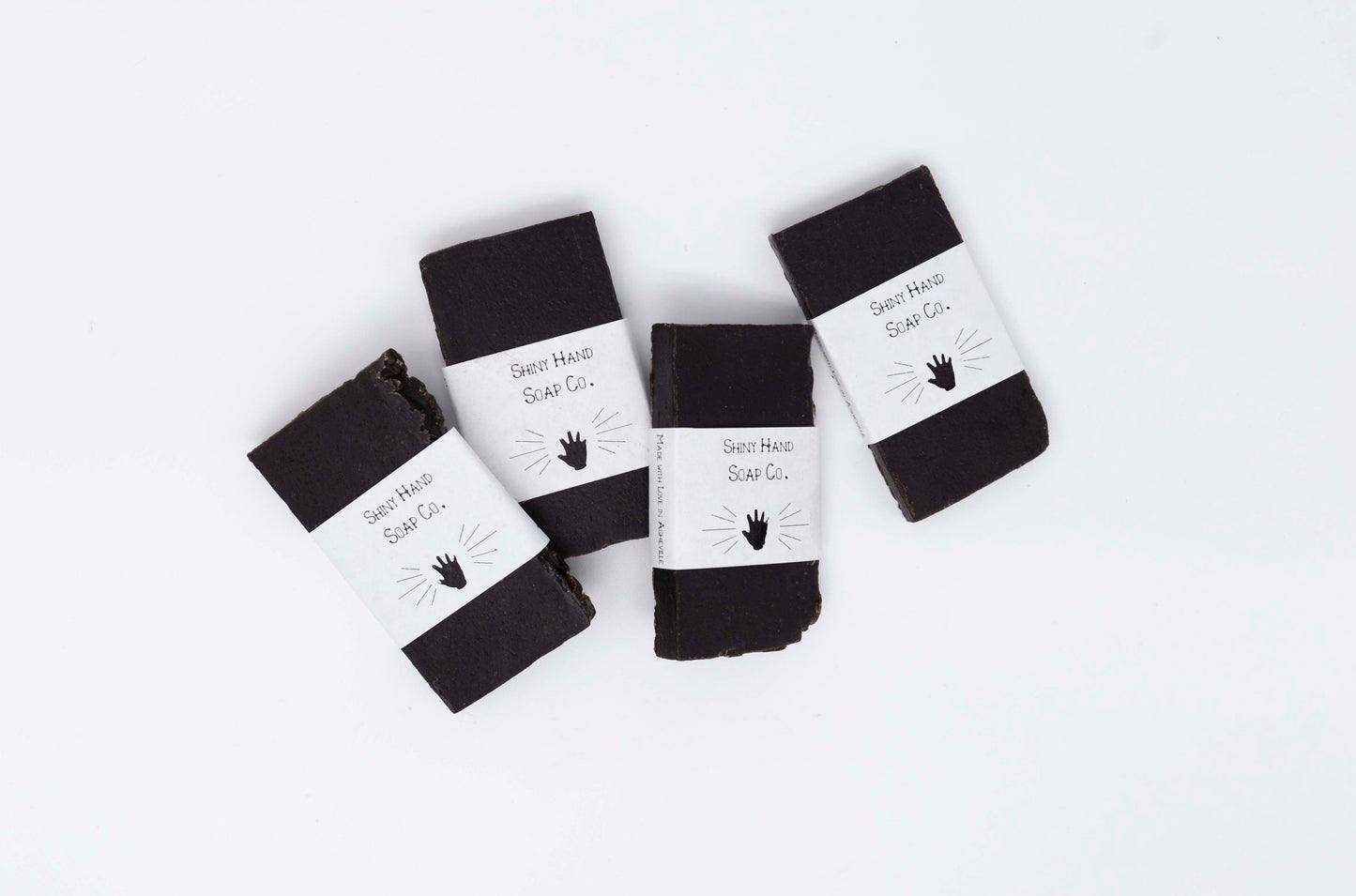 Four charcoal black midnight anise soap samples wrapped in crisp white recycled paper wrappers with a hand shaped cutout rest on a clean white backdrop.