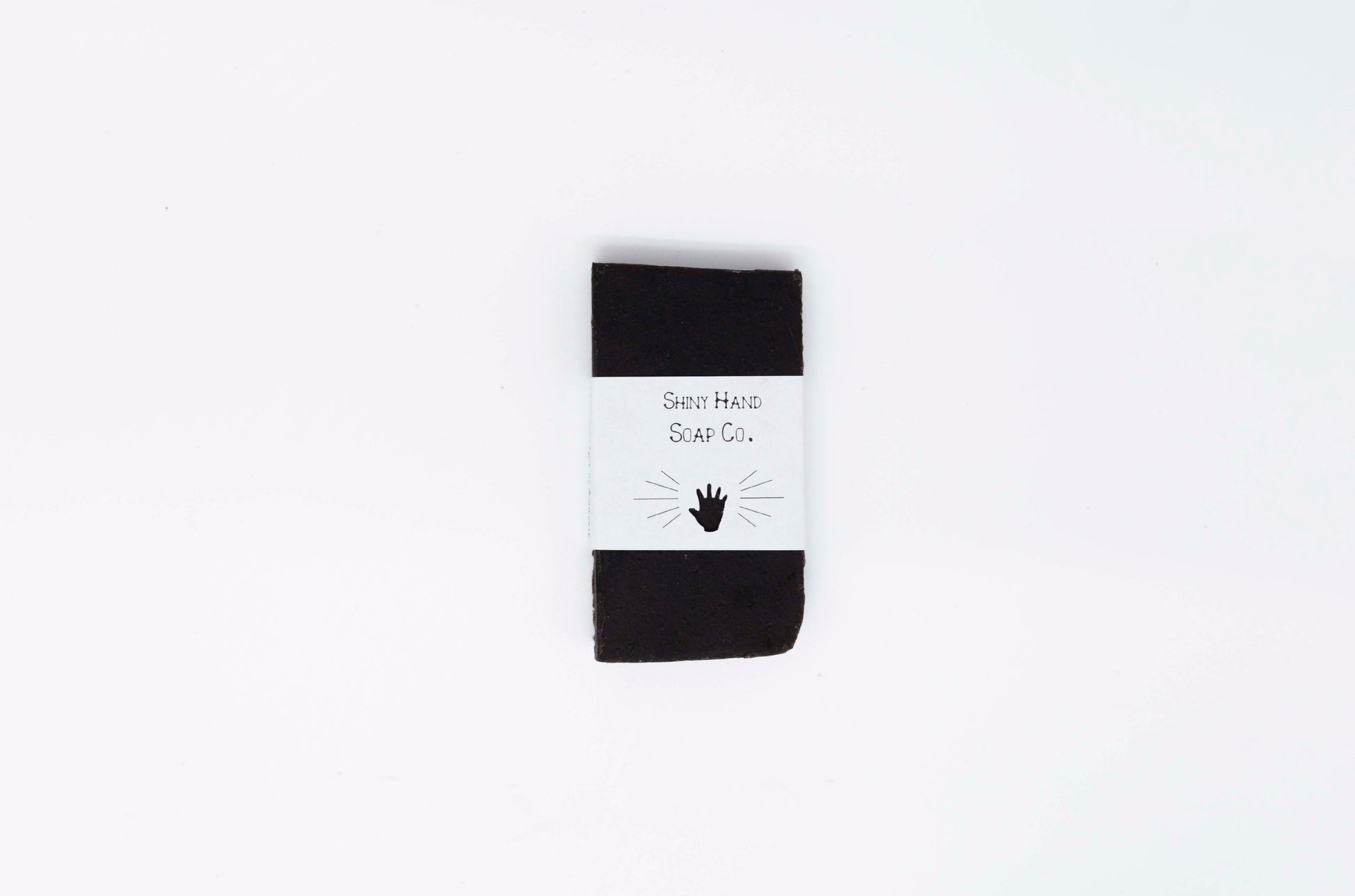 A charcoal black midnight anise soap sample wrapped in crisp white recycled paper wrapper with a hand shaped cutout rests on a clean white backdrop.