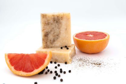 Ivory colored grapefruit black pepper soap bars with tiny specks of a black pepper swirl sit on a clean white background with fresh pink grapefruit slices, small black round peppercorns, and a sprinkling of crushed black pepper flakes.