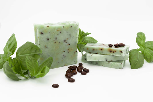 Pale Green Basil Mint Soaps flecked with bits of crushed coffee beans in front of a clean white backdrop with fresh green basil, fresh green mint leaves, and rich dark coffee beans.