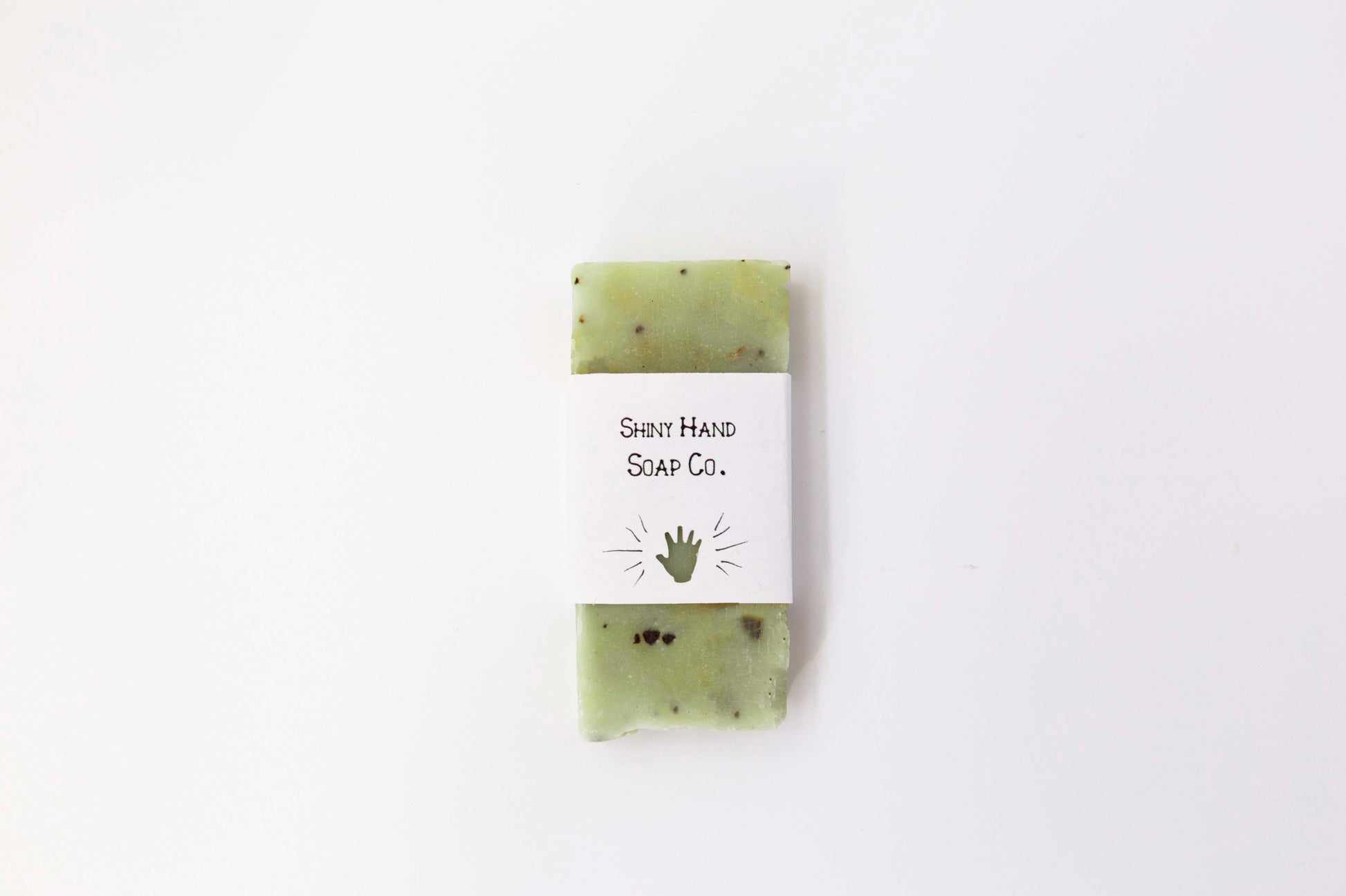 A miniature pale green basil mint soap flecked with crushed coffee beans sits on a clean white background wrapped in a small paper wrapper with a hand cut out of it.
