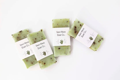 Four miniature pale green basil mint soaps flecked with crushed coffee beans sit on a clean white background with white paper wrappers that have a hand shape cut out of them.