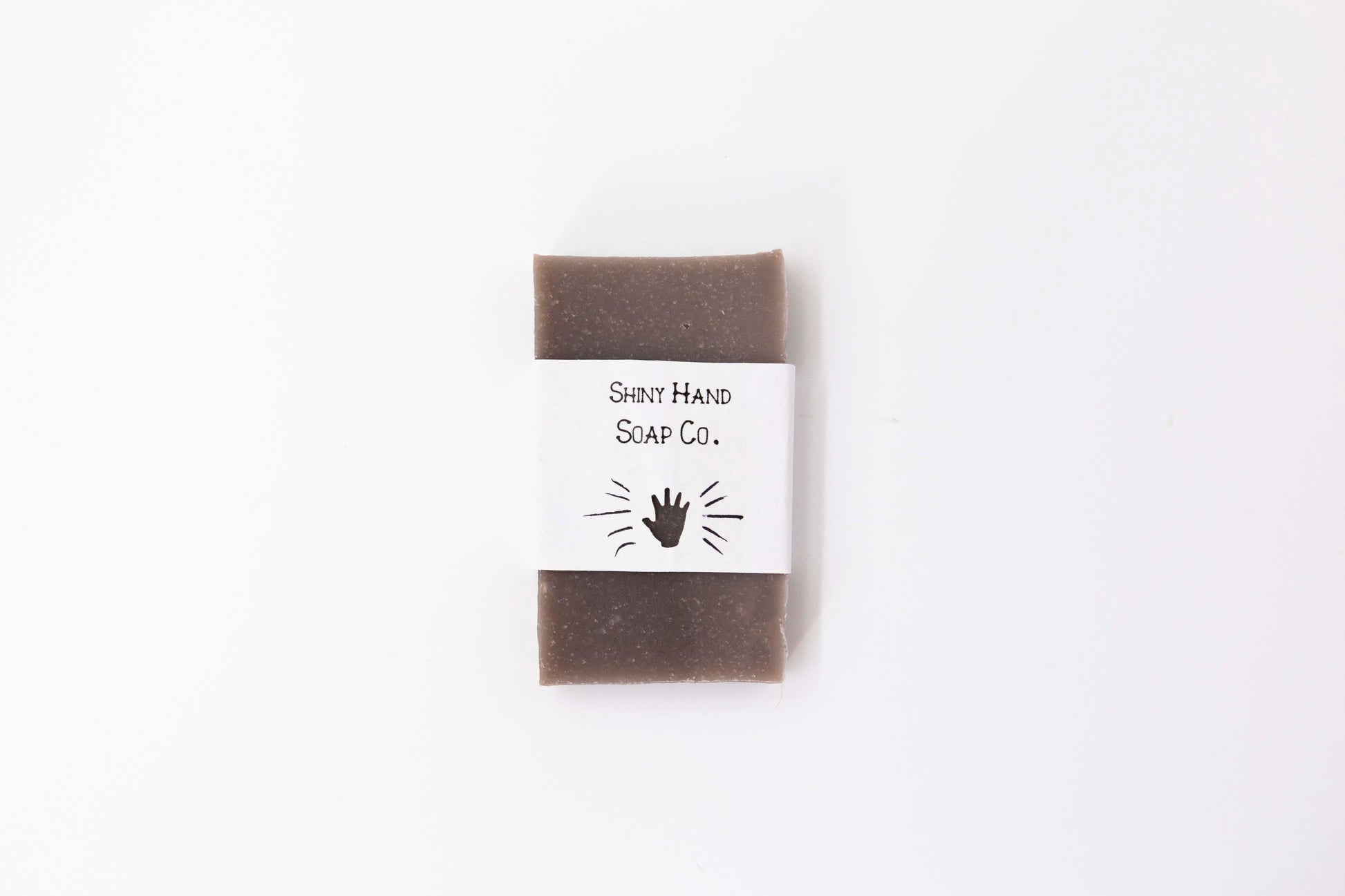 A brown cedarwood patchouli miniature bar soap sits on a clean white background wrapped in a small paper wrapper with a hand cut out of it.