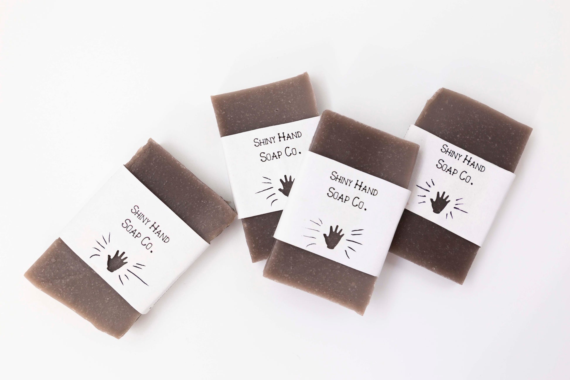 Four miniature brown cedarwood patchouli bar soaps sit on a clean white background with white paper wrappers that have a hand shape cut out of them.