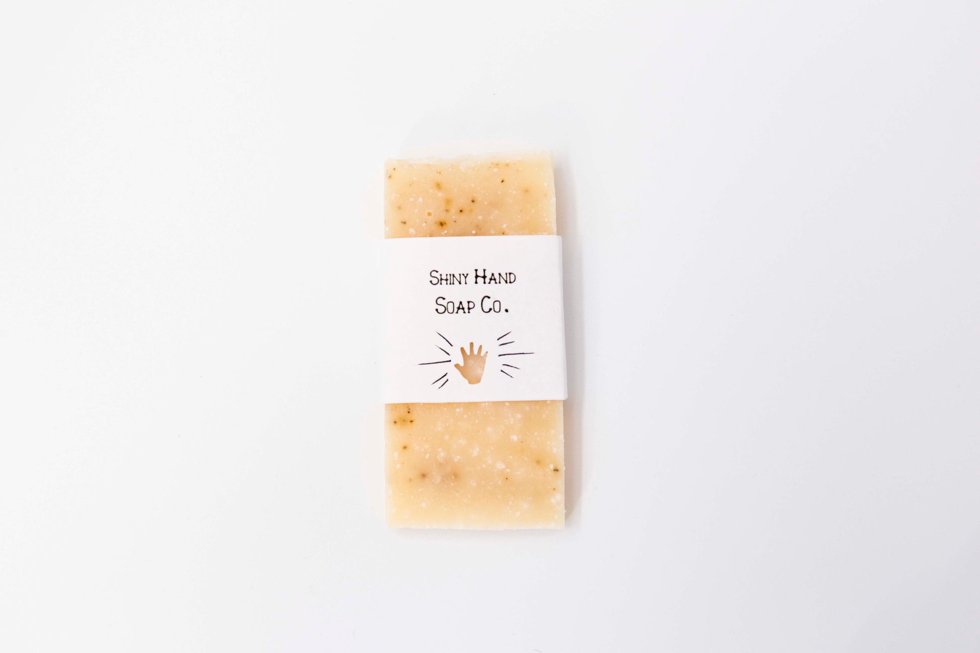 A miniature ivory colored grapefruit black pepper soaps with tiny specks of a black pepper swirl sits on a clean white background wrapped in a small paper wrapper with a hand cut out of it.