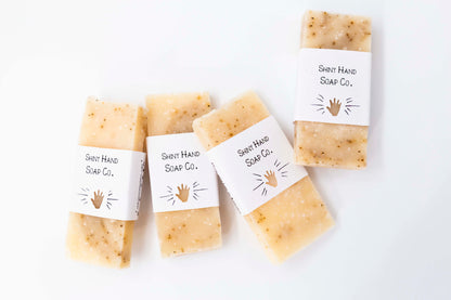 Four miniature ivory colored grapefruit black pepper soaps with tiny specks of a black pepper swirl sit on a clean white background with white paper wrappers that have a hand shape cut out of them.