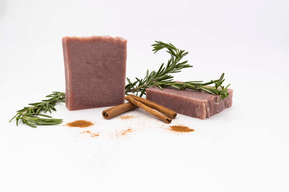 Pale red Cinnamon rosemary soap bars speckled with dark brown cinnamon sit on a clean white background surrounded by fresh green rosemary springs, orange powdered cinnamon, and curls or real cinnamon bark
