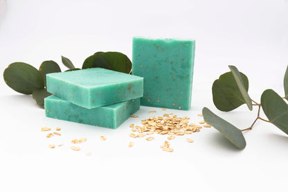 Aqua blue eucalyptus oatmeal soaps flecked with ivory colored oats sit on a clean white background with fresh pale green and blue eucalyptus leaves and organic light tan oats.
