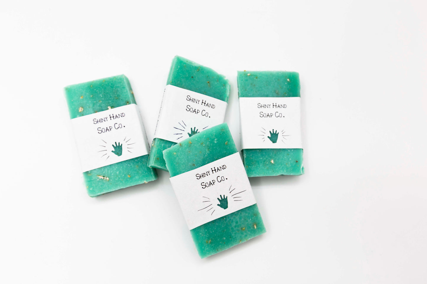 Four miniature aqua blue eucalyptus oatmeal soaps flecked with ivory colored oats sit on a clean white background with white paper wrappers that have a hand shape cut out of them.